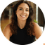 Smiling woman with long brown curly hair wearing a black ribbed tank top indoors.