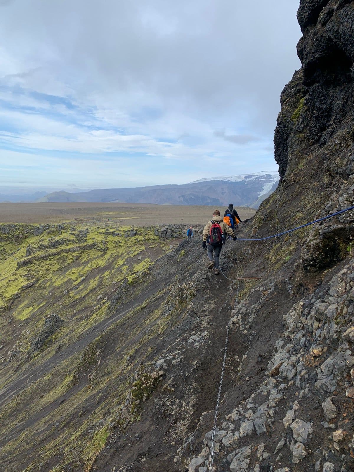Hikers trekking along a narrow, rocky path with a safety chain on the Fimmvörðuháls trail in Iceland.