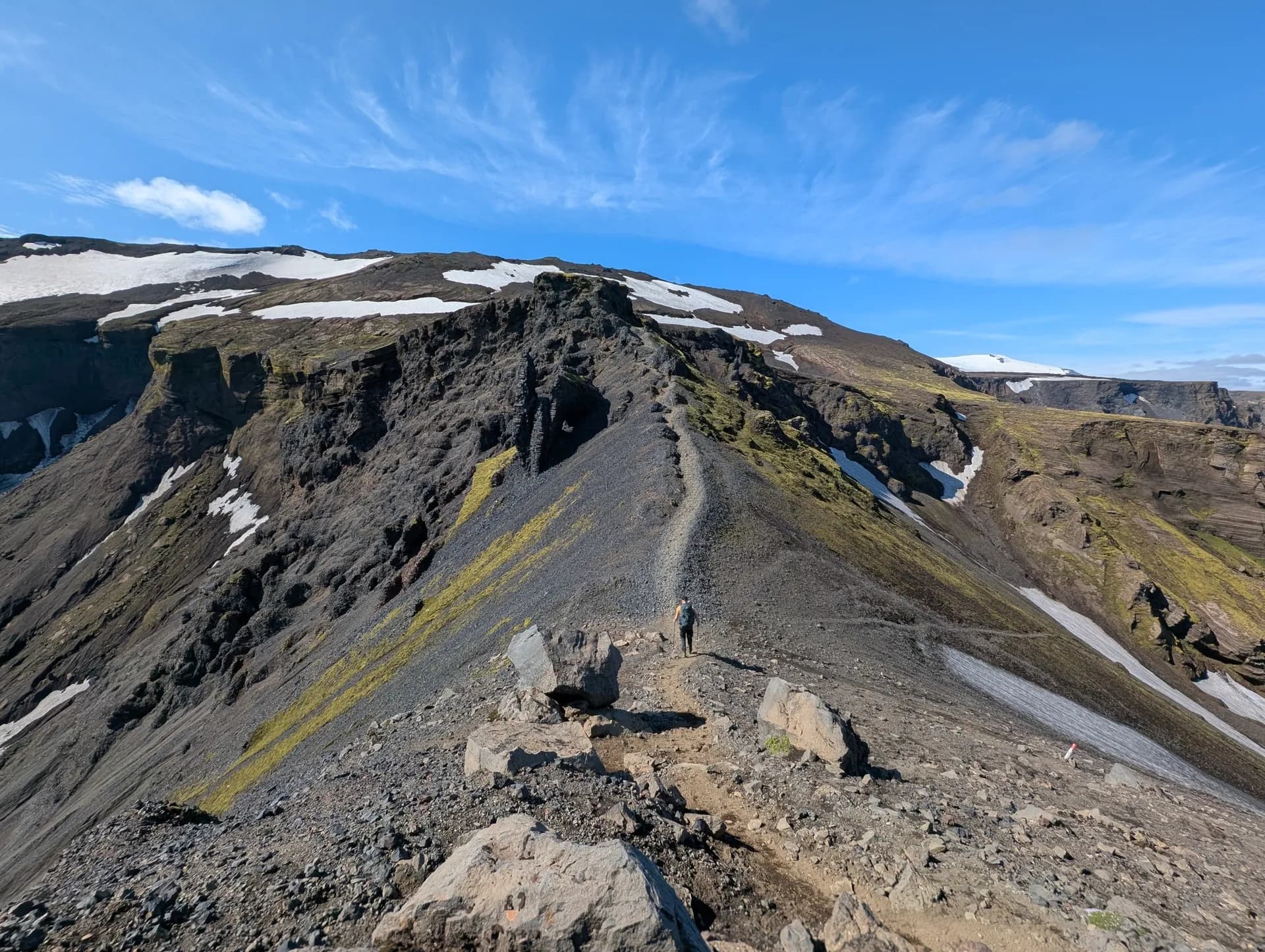 Hiker on the Fimmvörðuháls trail along a rocky mountain ridge with patches of snow under a blue sky.