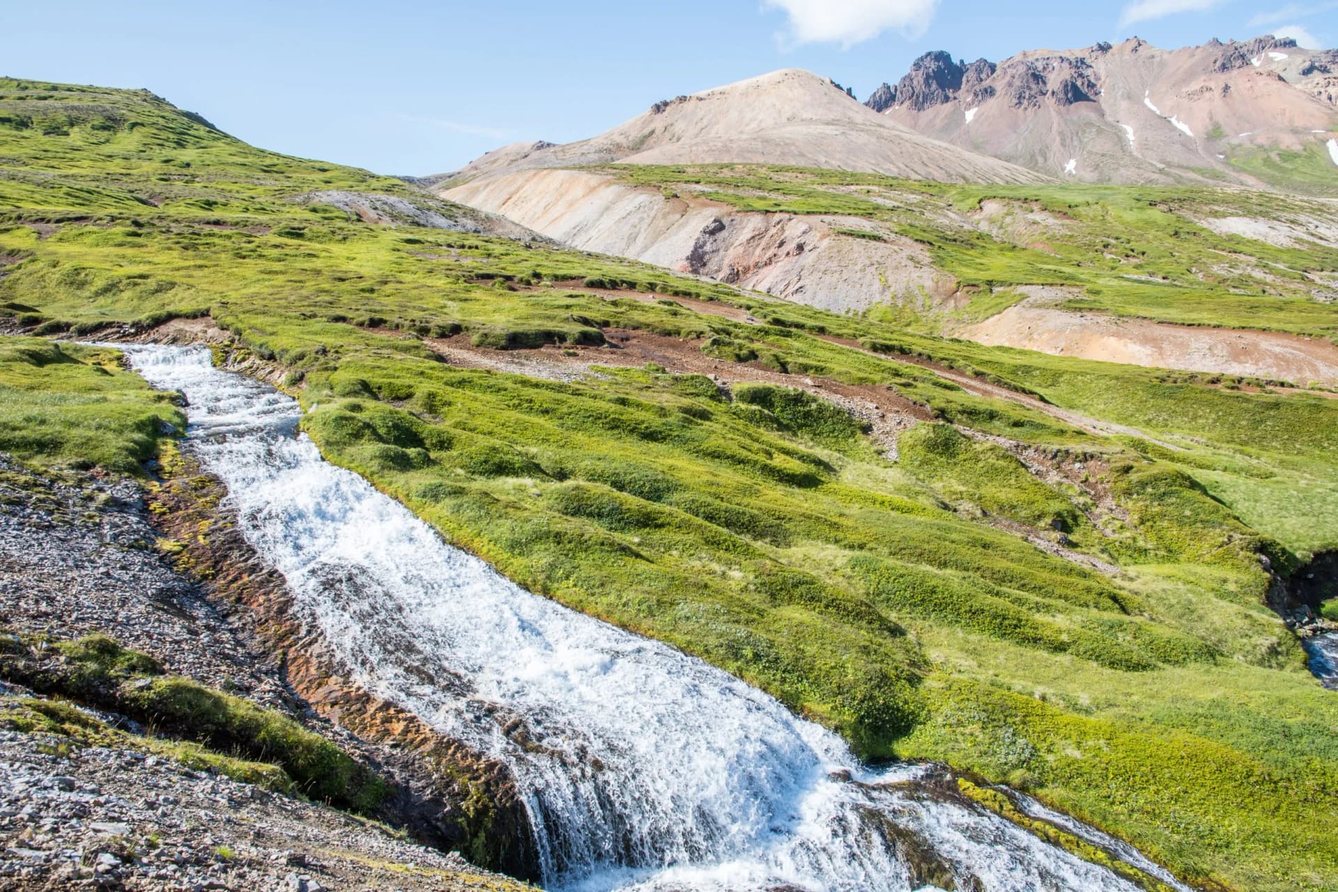 Cascading river rapids flowing down a bright green, grassy hillside toward mountains in Breidavik, Iceland.