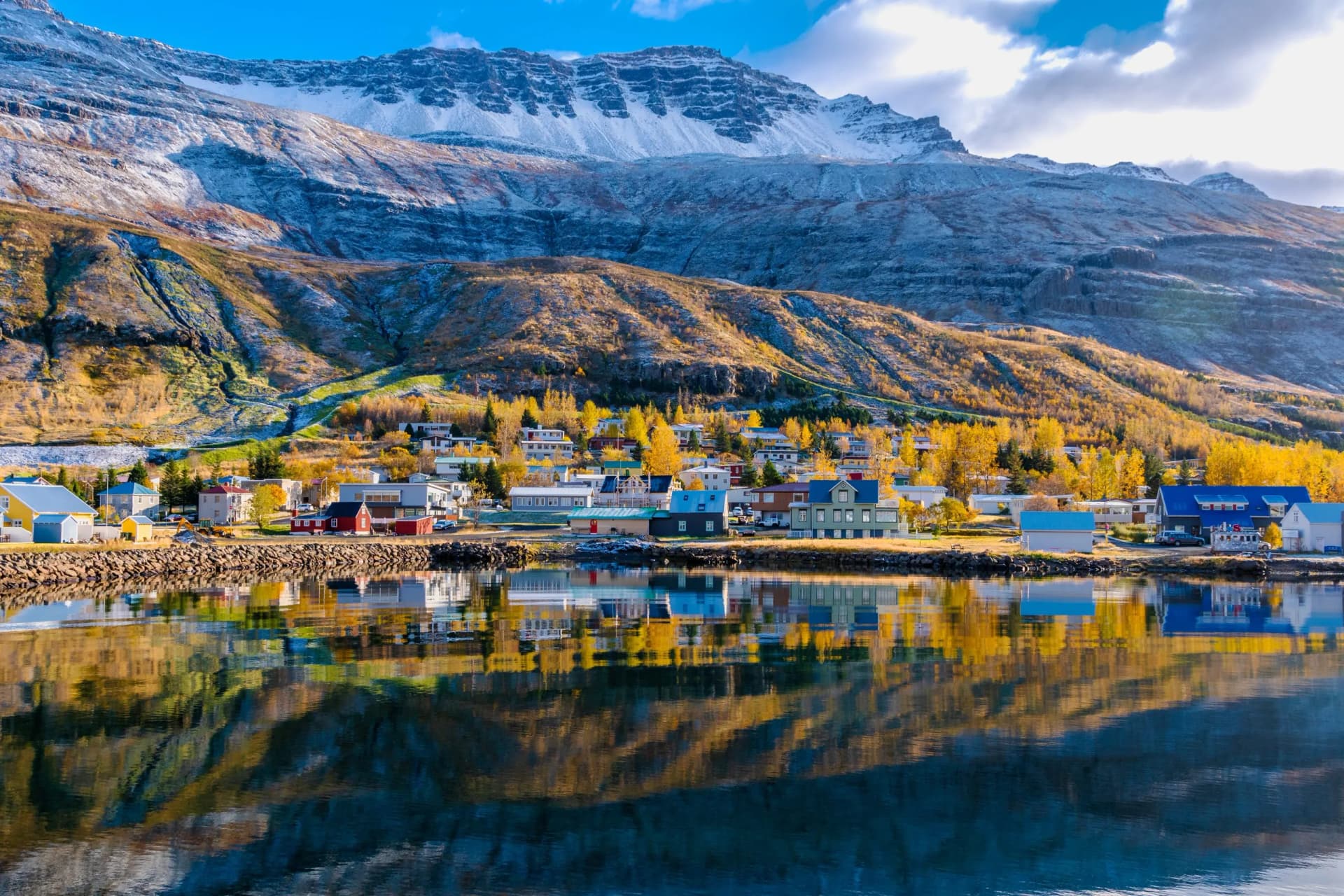 Seydisfjordur town reflected in water with snow-dusted mountains and autumn trees.