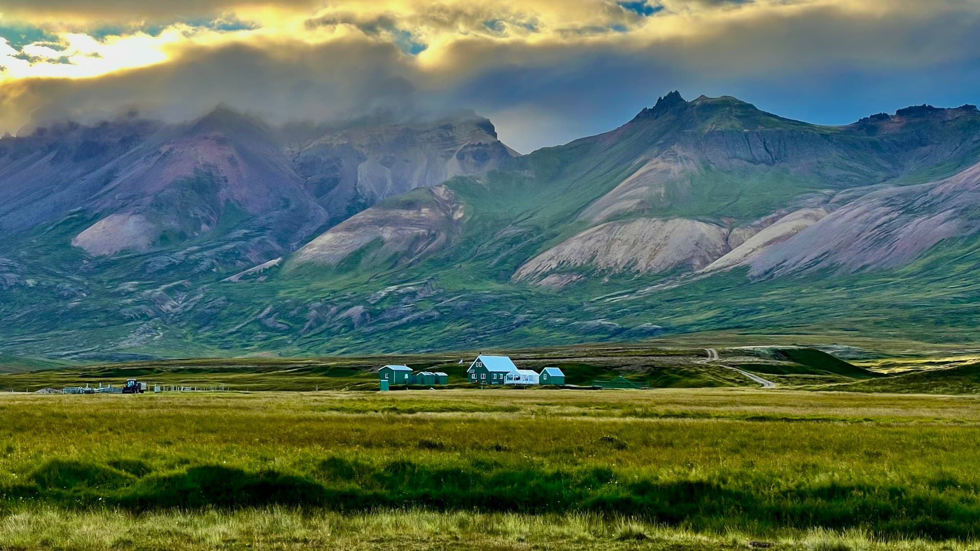 Colorful mountains above a green plain with small buildings under a cloudy sky