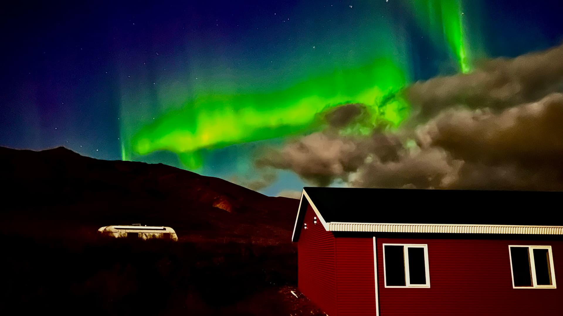 Northern lights glowing in the night sky over a red hut and dark mountains