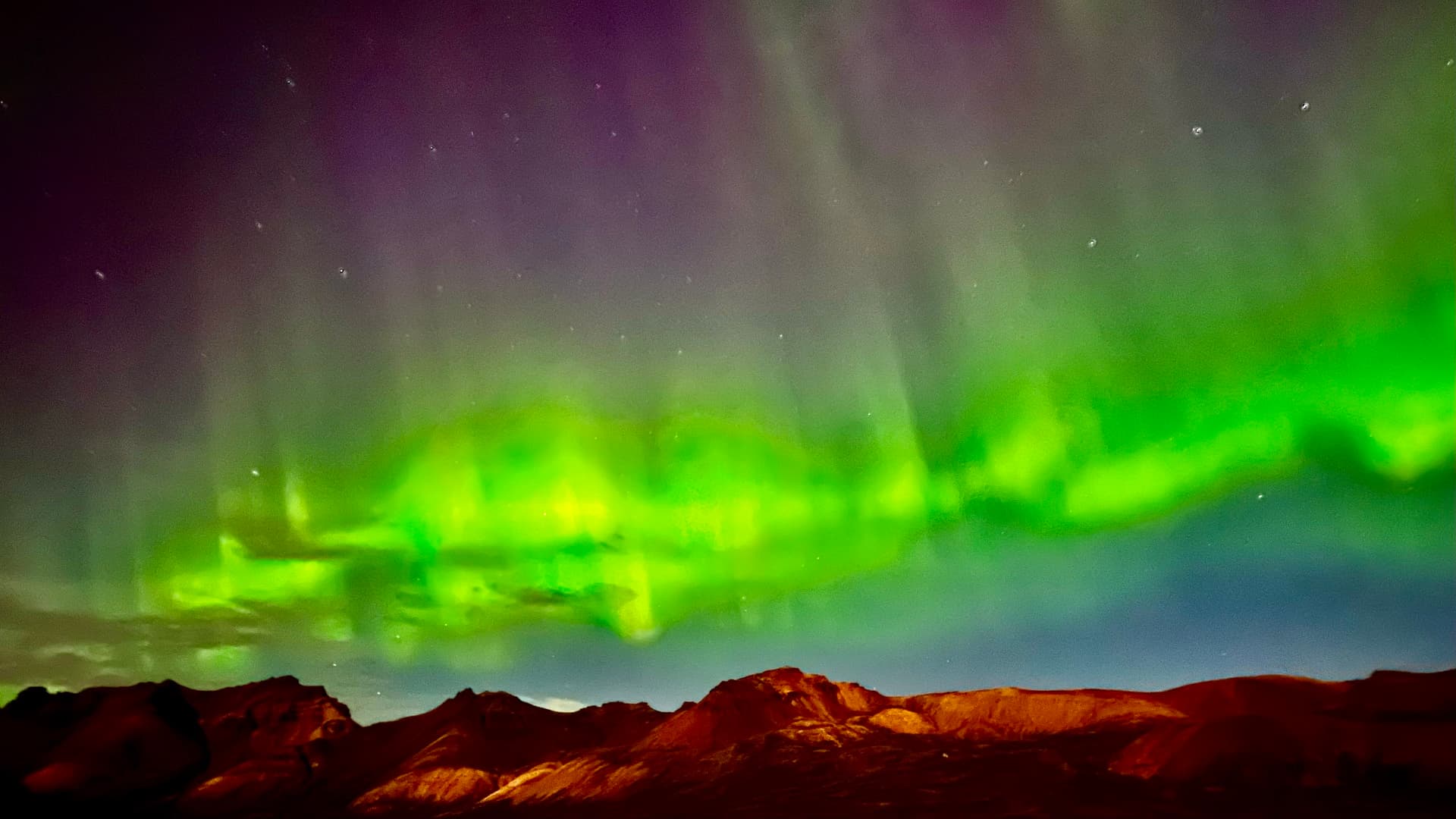 Northern lights glow green and purple over dark mountain ridges at night