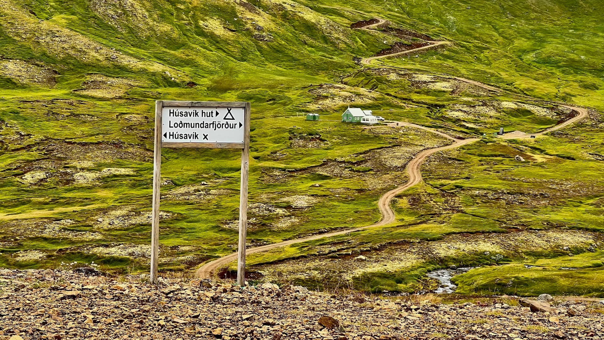 Wooden hiking sign and zigzag dirt path winding up green mountain slopes