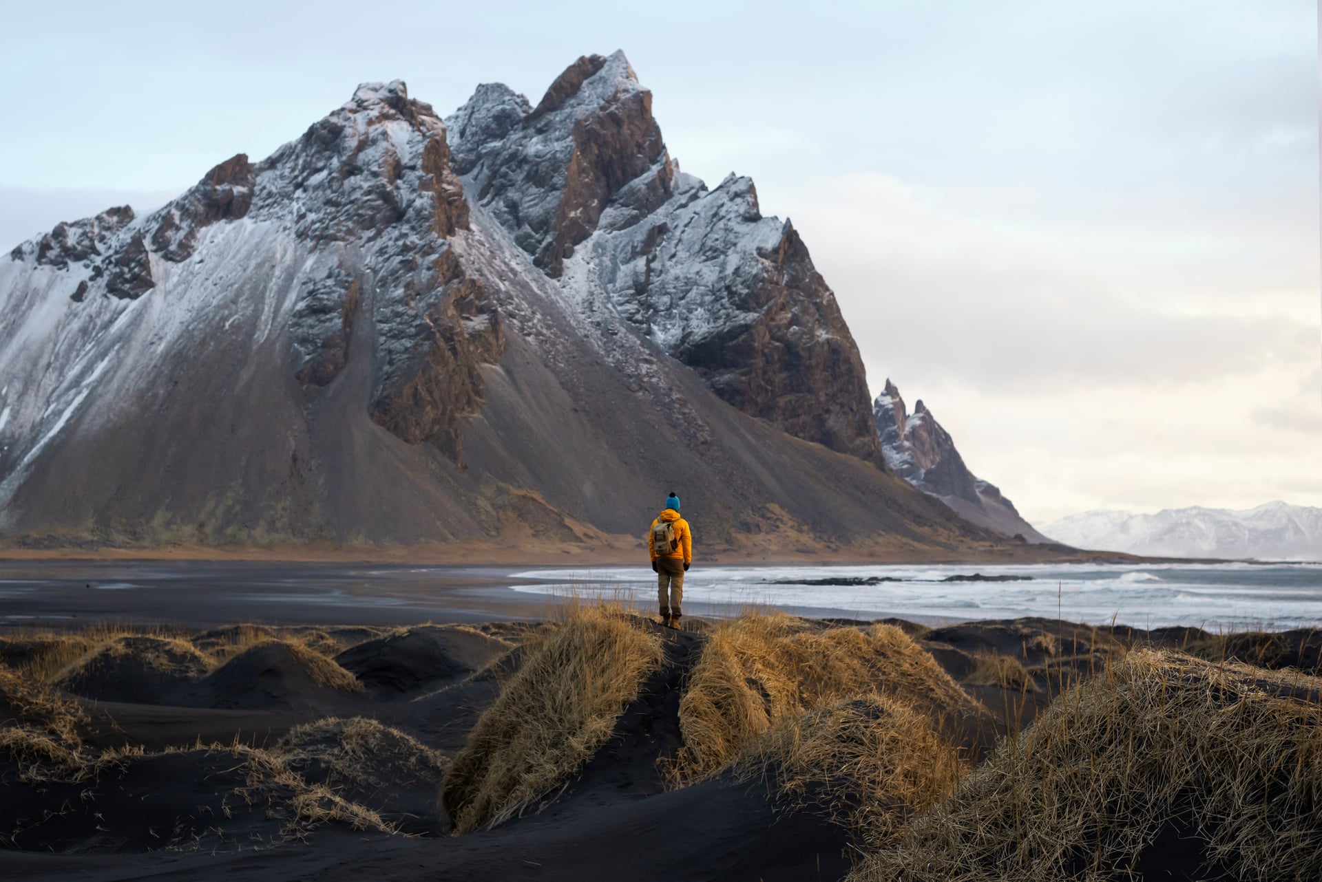 Hiker overlooking black sand dunes, ocean, and snow-dusted mountains in Iceland.