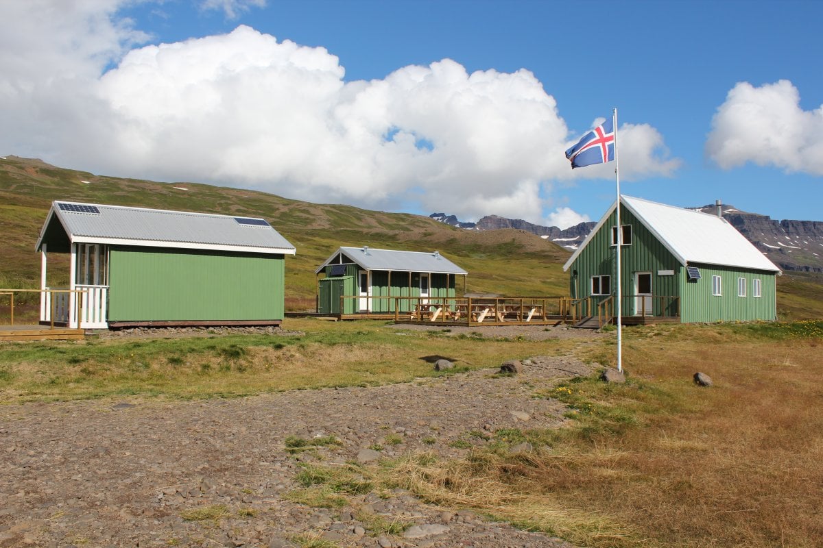 Grønne hytter med solpaneler og et islandsk flag nær bjerge i Loðmundarfjörður.