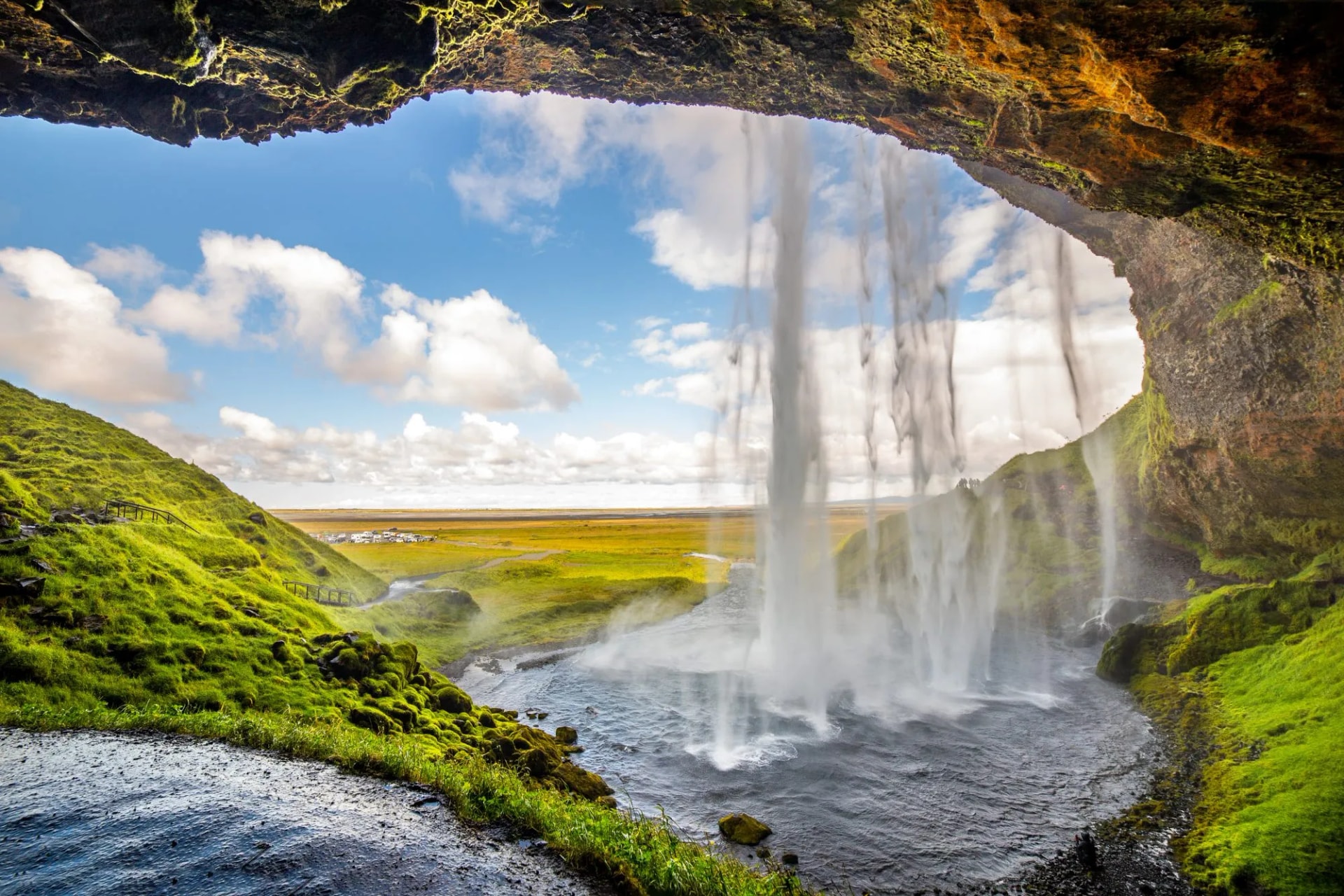 View from behind Seljalandsfoss waterfall in Iceland with green mossy cliffs and blue sky.
