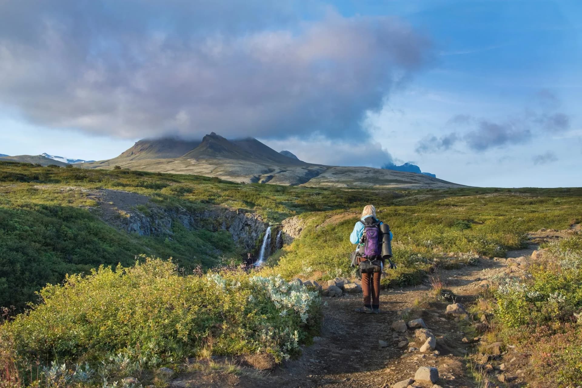 Hiker with backpack on dirt trail approaching waterfall in Skaftafell National Park, Iceland.