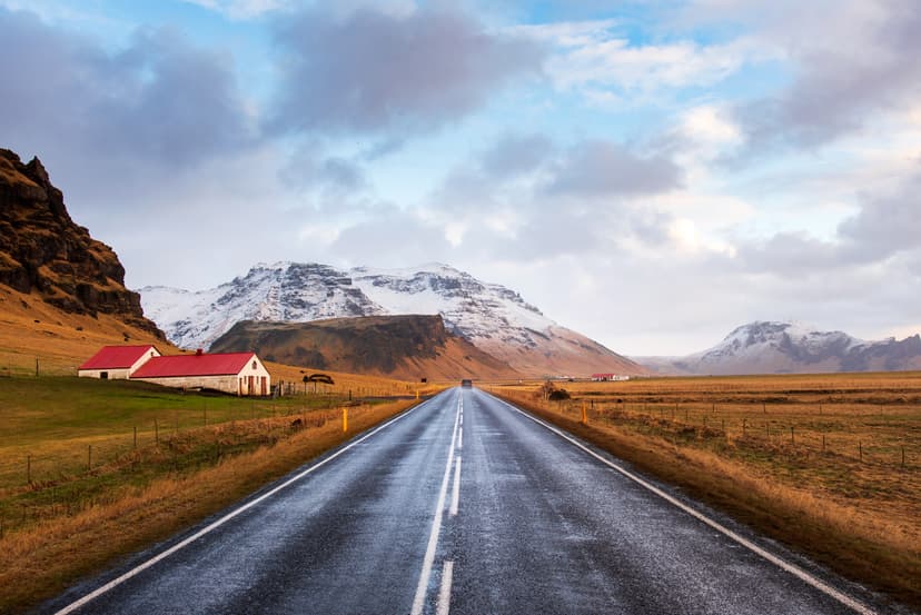 Asphalt road stretching through a rural landscape with snow-capped mountains and a building with a red roof.