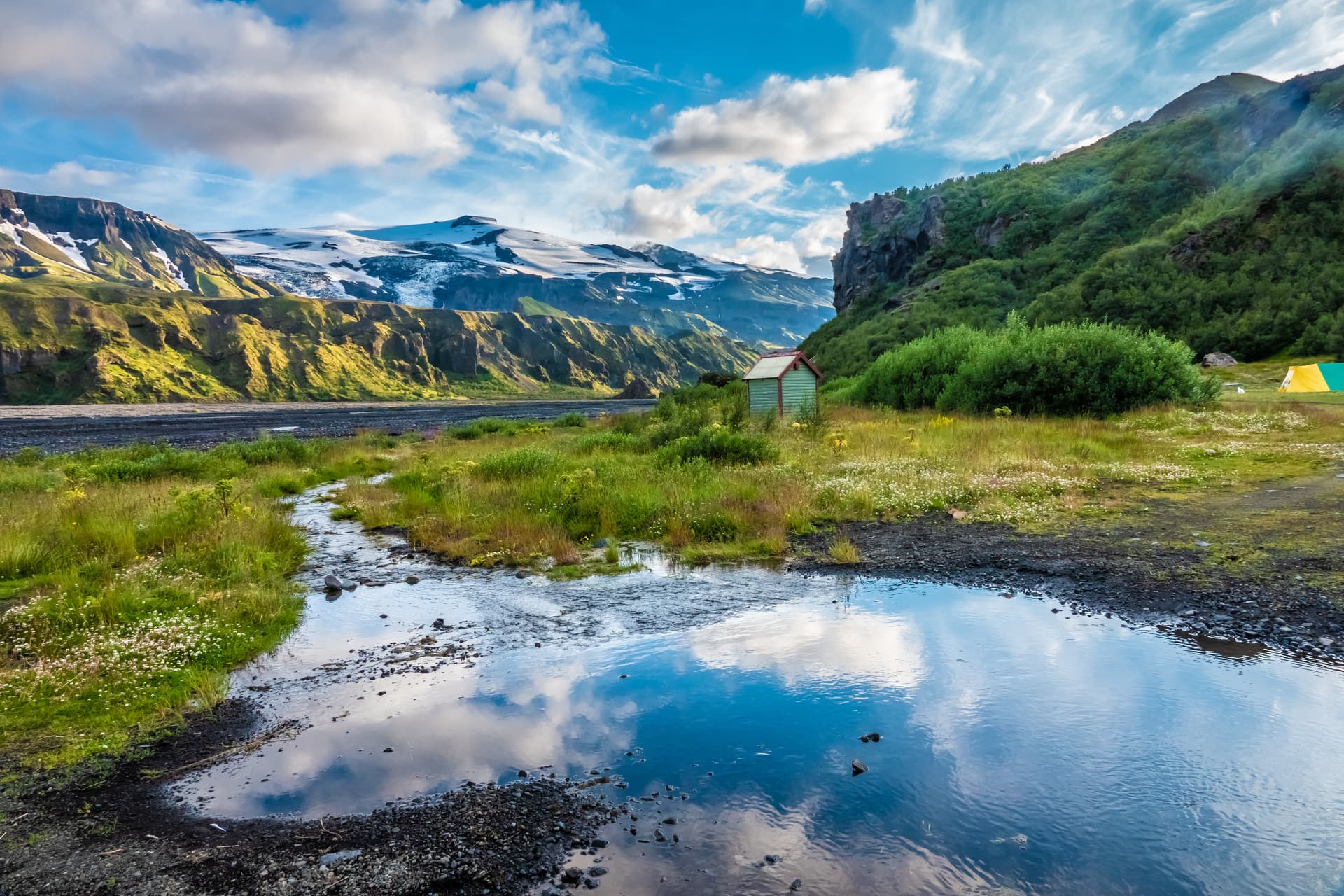 Thórsmörk landscape with glacial mountains, stream reflection, green hills, and small hut.