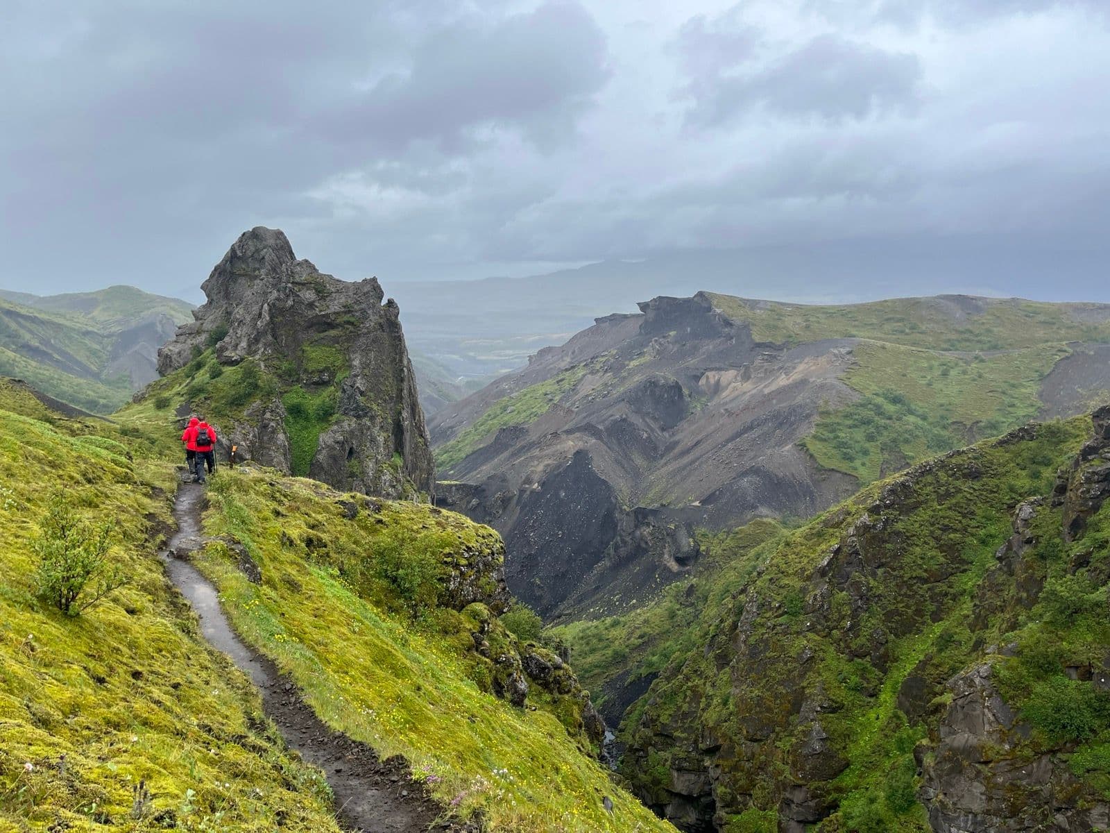 Hikers on muddy trail above mossy canyon with dramatic rock formations under cloudy sky.