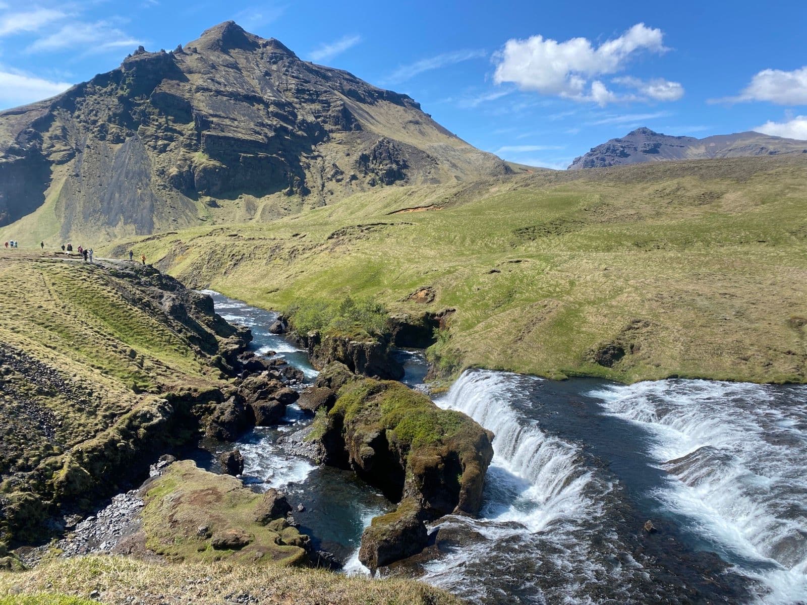 River cascading over mossy rocks near Fimmvörðuháls, with hikers on green hillside under blue sky.