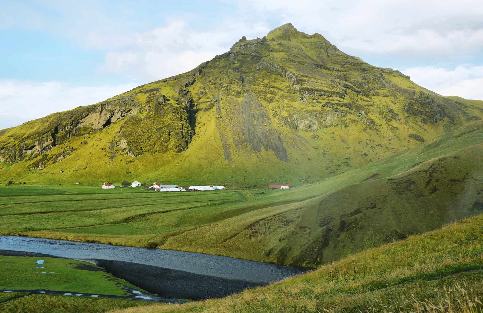 Mountain covered in green moss overlooking a river, farm buildings, and green fields at Skógafoss.