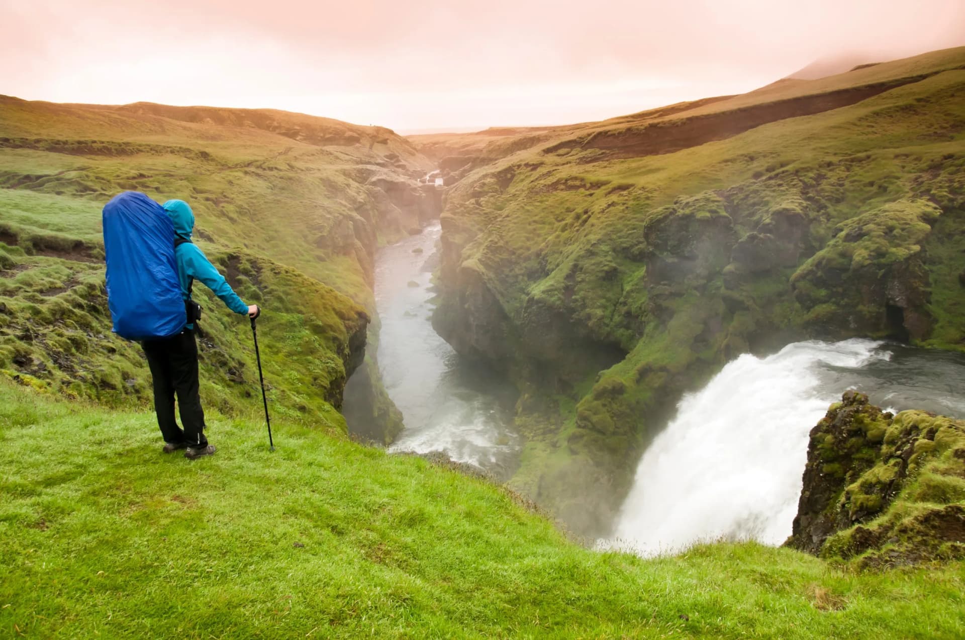 Hiker with blue backpack overlooks Skógafoss waterfall canyon on Laugavegur Trail.