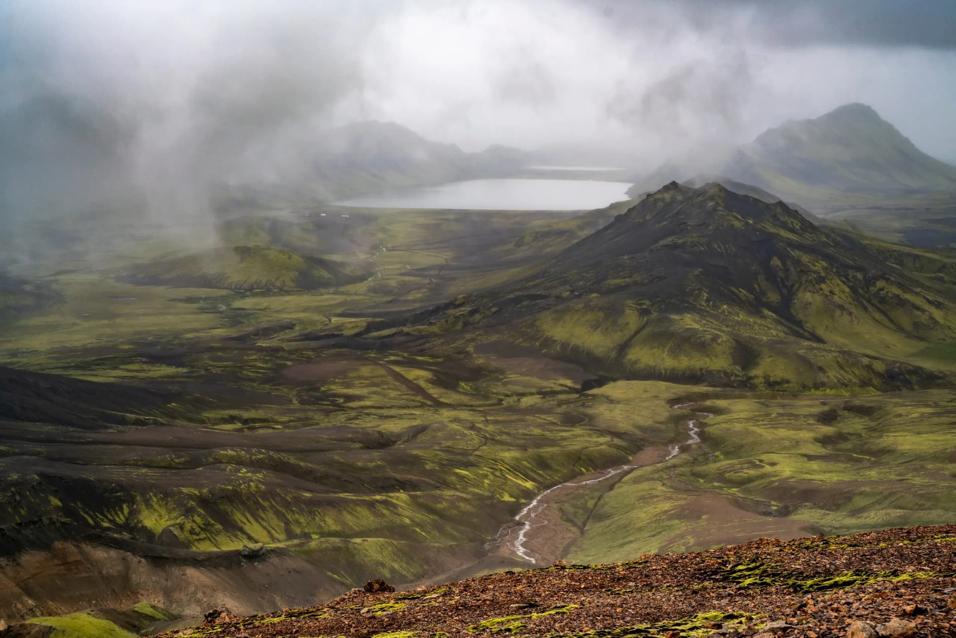 Mountain landscape with green mossy slopes, winding river, and lake under heavy fog near Alftavatn.