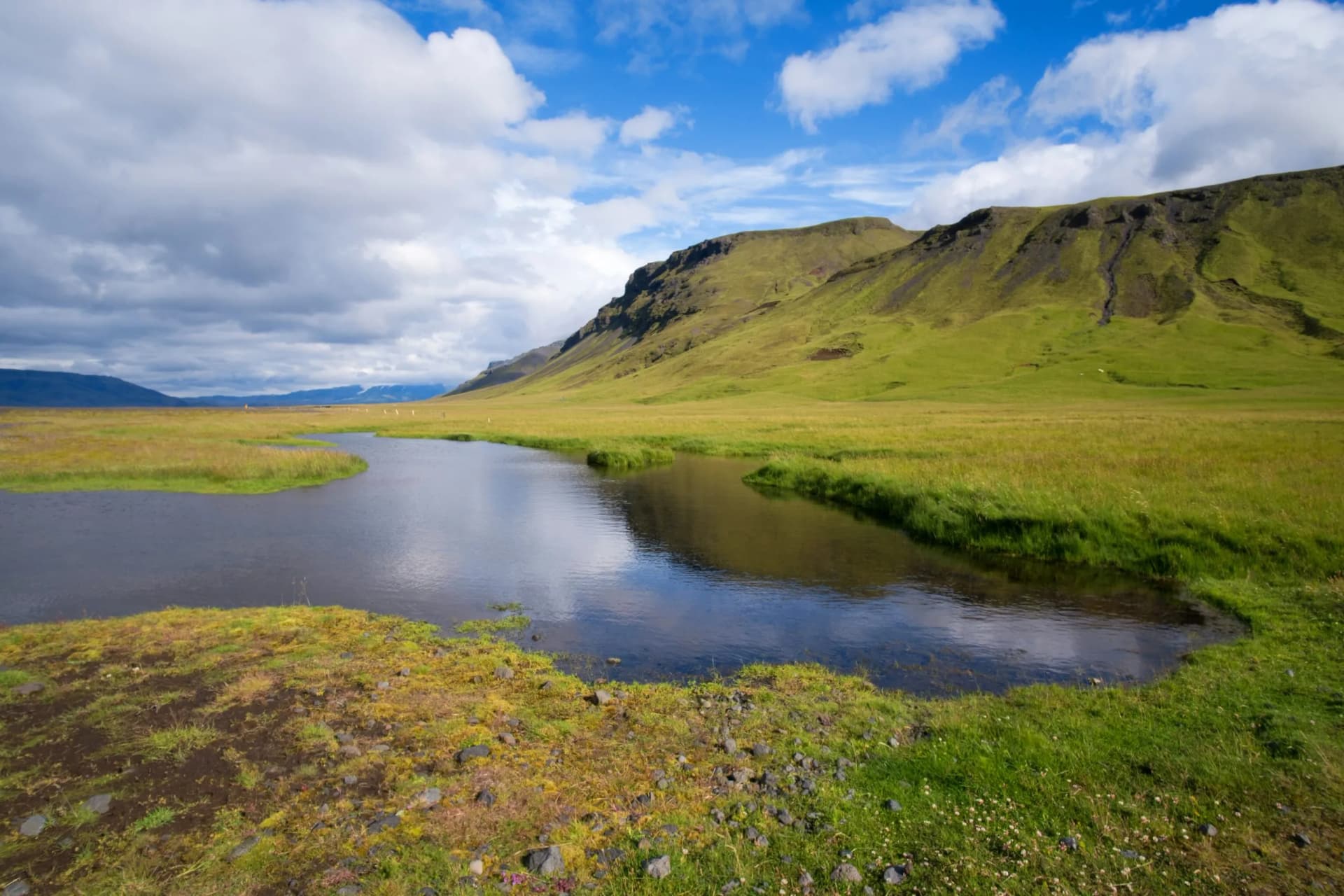 Winding stream through green grassy plain beneath large, steep green mountains under a cloudy blue sky.