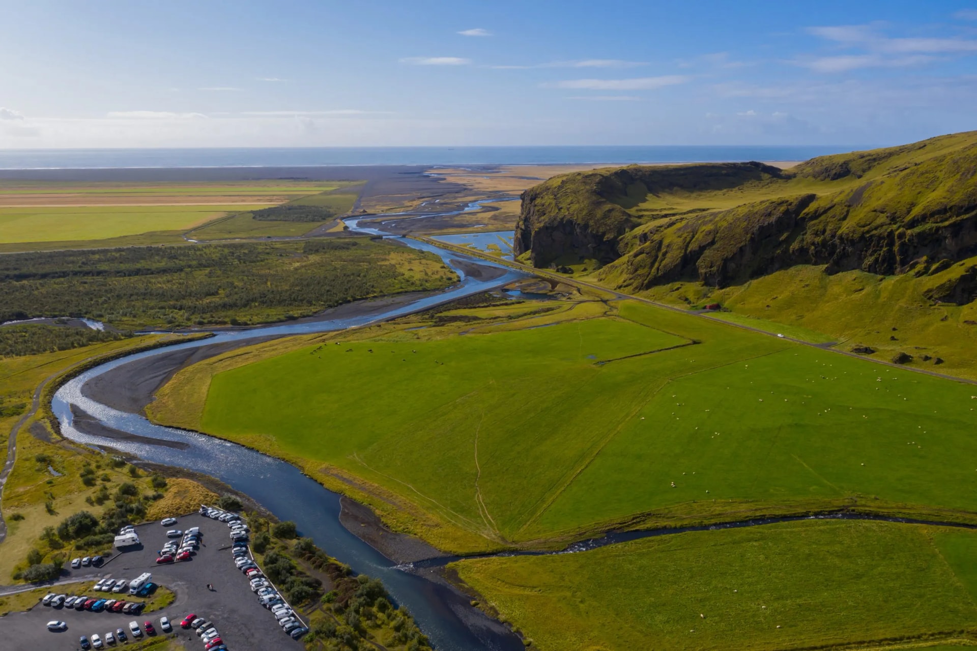 Lush green Skoga River valley near Skogafoss waterfall with parking lot and ocean view.