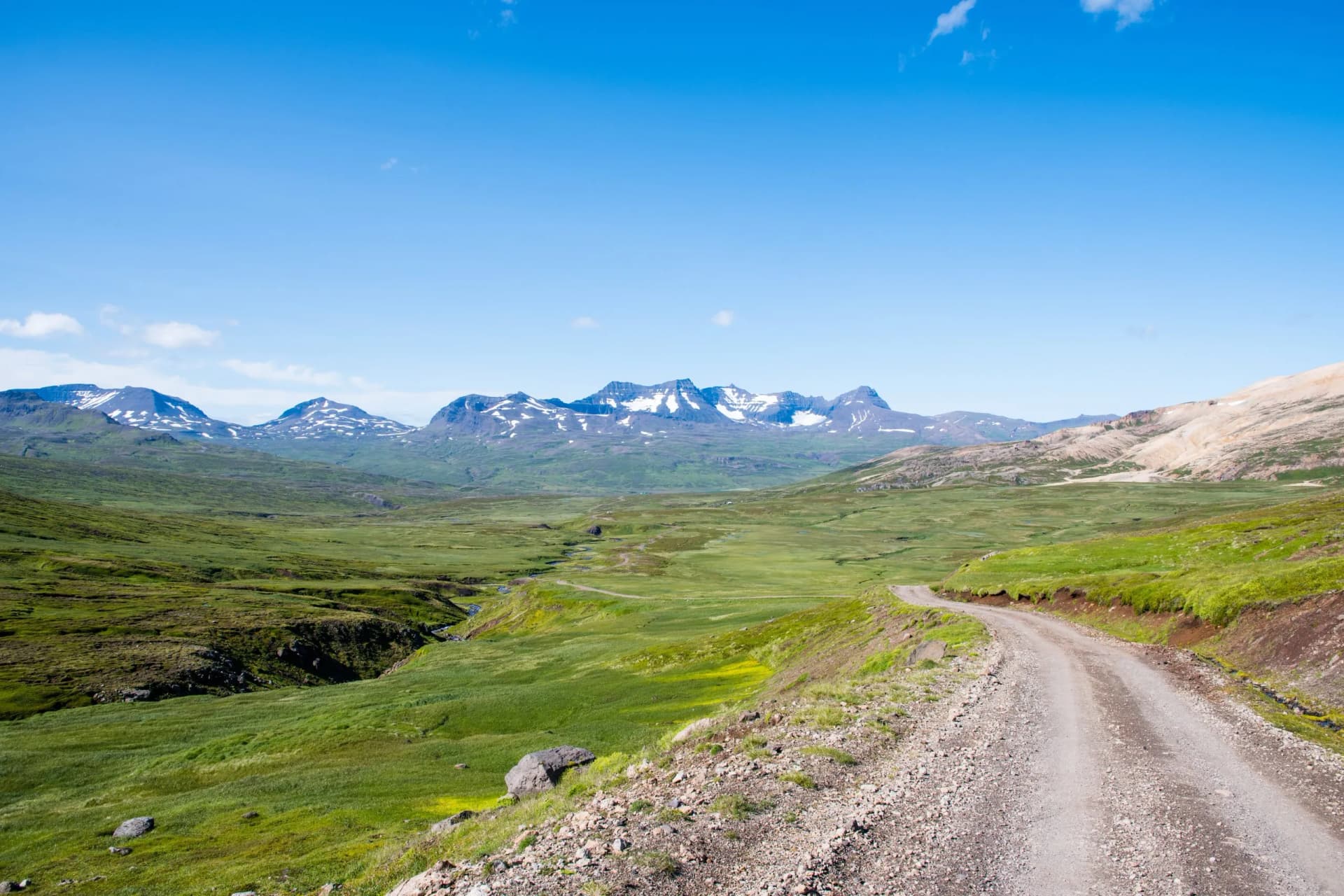 Gravel road winding through green valley toward snow-capped mountains in Borgarfjordur Eystri, Iceland.